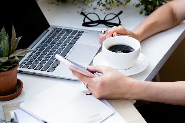 Die Hand der Frau hält eine Tasse Kaffee und ein Smartphone in einem gemütlichen Büro mit Laptop und grünen Pflanzen