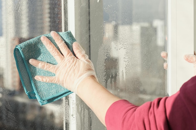 Die Hand der Frau in einem Gummihandschuh wischt Fenster mit einem Lappen ab