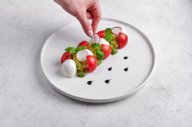 Die Hand der Frau legt die Radieschenscheibe in einen köstlichen italienischen Caprese-Salat mit reifen Tomaten, frischem Gartenbasilikum und Mozzarella-Käse