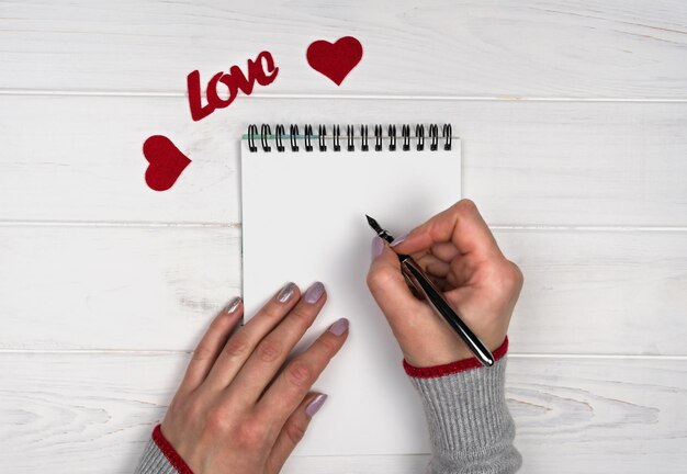 Die Hand der Frau schreibt eine Liebesbotschaft auf einen Notizblock Flatlay-Komposition Liebesromantik für den Valentinstag Valentinstag-Ferienkonzept Top-View