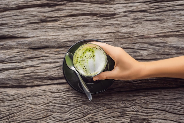 Die Hand der Frauen hält eine Tasse Matcha Latte, grüner Tee, auf einem alten hölzernen Hintergrundtisch.