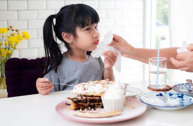 Die Hand der Mutter benutzt ein Tuch, um den Mund ihrer asiatischen Tochter von Lebensmittelflecken zu reinigen, während sie am Tisch süßen Kuchen im Café isst.