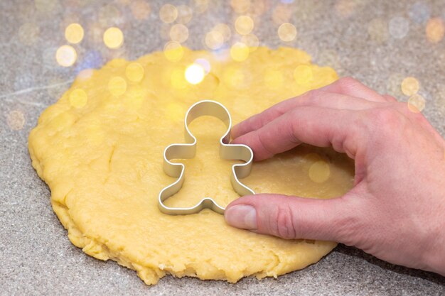 Die Hand des Küchenchefs schneidet die Form von Lebkuchenmännern aus rohem Rollteig auf einem fabelhaften Hintergrund mit gelbem Bokeh. Der Prozess der Herstellung von Weihnachtsplätzchen zu Hause. Weihnachtsessentradition