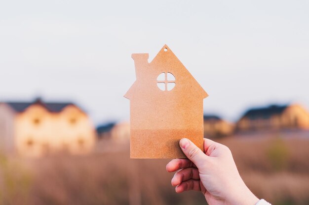 Die Hand des Mannes hält ein Holzhaus auf dem Hintergrund von verschwommenen Gebäuden