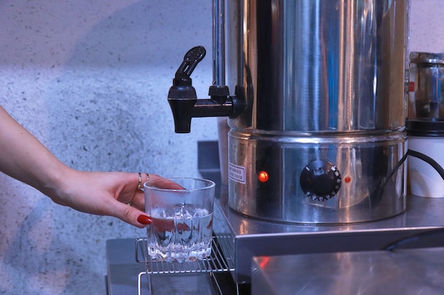 Die Hand einer Frau drückt auf den Wasserhahn, um heißes Wasser aus dem Kessel in ein klares Glas auf dem Tisch zu gießen