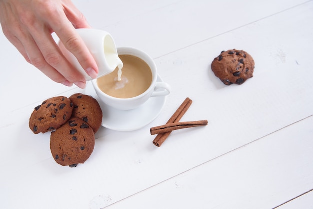 Die Hand einer Frau gießt Milch in Kaffee. Eine Tasse duftenden Kaffees mit Zimt und Keksen auf einem weißen Hintergrund.