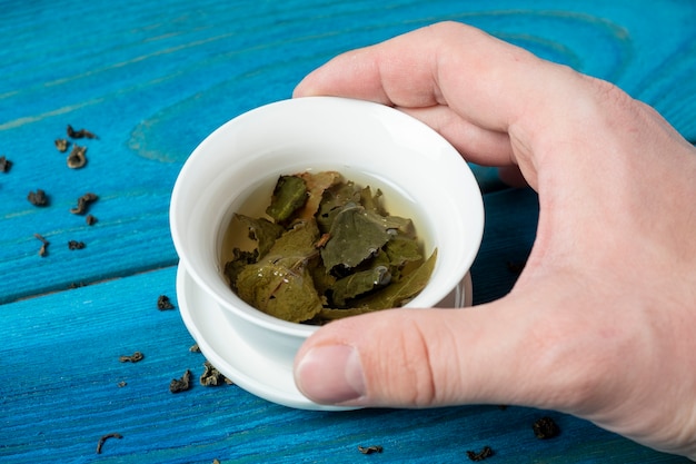 Die Hand einer Frau hält eine weiße Porzellanschale mit Tee. Blauer hölzerner Hintergrund. Die geöffneten Blätter des großblättrigen Tees. Chinesischer Tee. Tee-Party. Ansicht von oben, flach