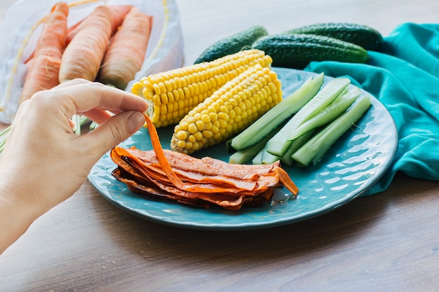 Die Hand einer Frau nimmt ein Stück gekochten Karottenspeck auf einem türkisfarbenen Teller mit Mais und Gurke. Vegetarisches Essen, Fleischersatz, Protein, gesunder Snack