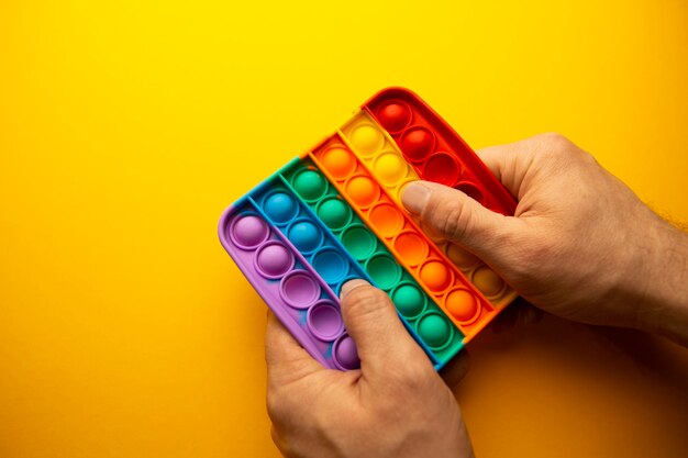 Die Hand eines Mannes drückt auf ein Regenbogen-Silikon-Antistress-Spielzeug, oder ein einfaches Grübchen auf einem gelben Hintergrundkopierraum