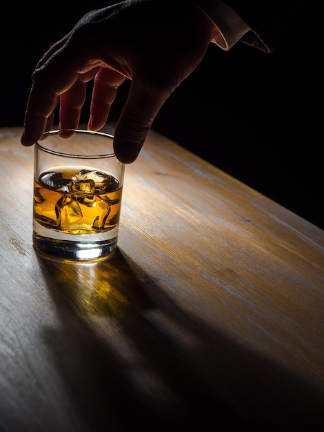 Die Hand eines Mannes hält ein Glas Whiskey auf einem Holztisch in einem dunklen Raum