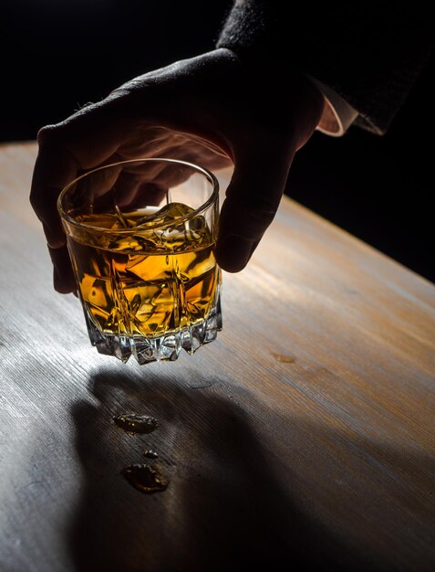 Die Hand eines Mannes hält ein Glas Whiskey über einem Tisch in einem dunklen Raum