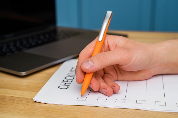 Die Hand eines Mannes hält einen Stift und schreibt eine Checkliste mit Kontrollkästchen, einen Holztisch