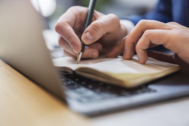 Die Hand eines Mannes in Nahaufnahme beim Schreiben in einem Notizblock auf einem modernen Laptop vor einem Softfokus-Hintergrund