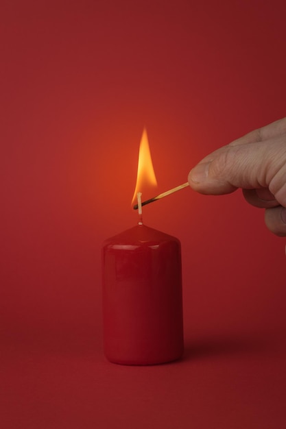 Die Hand eines Mannes mit einem brennenden Streichholz zündet eine rote Kerze auf rotem Grund an. Das Konzept einer romantischen Beziehung.