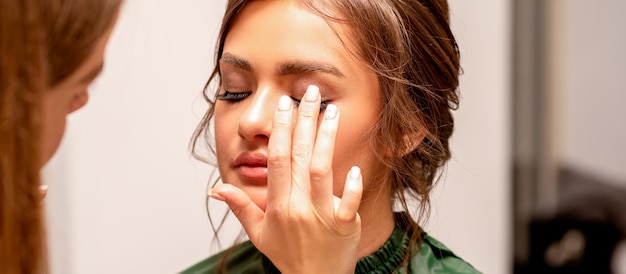 Die Hand eines Maskenbildners trägt in einem Schönheitssalon mit den Fingern Lidschatten auf das Augenlid einer jungen kaukasischen Frau auf