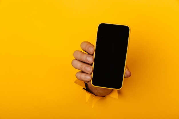 Die Hand hält ein Smartphone aus einem Loch, das in einer gelben Papierwand mit Kopierraum zerrissen ist