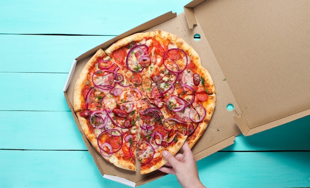Die Hand nimmt ein Stück Pizza in einer Schachtel auf einem blauen Betontisch. Draufsicht