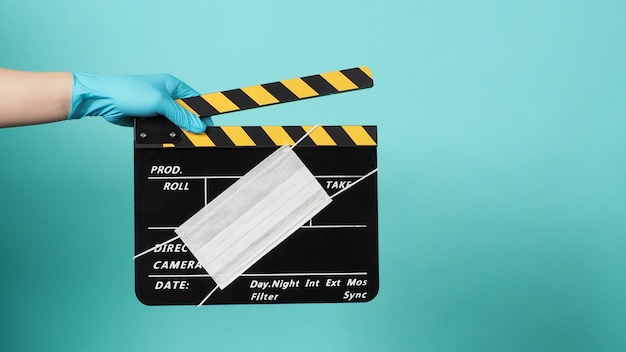 Die Hand trägt blaue medizinische Handschuhe, die schwarzes Clapper Board oder Schiefer mit Gesichtsmasken halten. es wird in der Filmproduktion und in der Kinoindustrie auf grünem oder blauem Tiffany-Hintergrund verwendet.