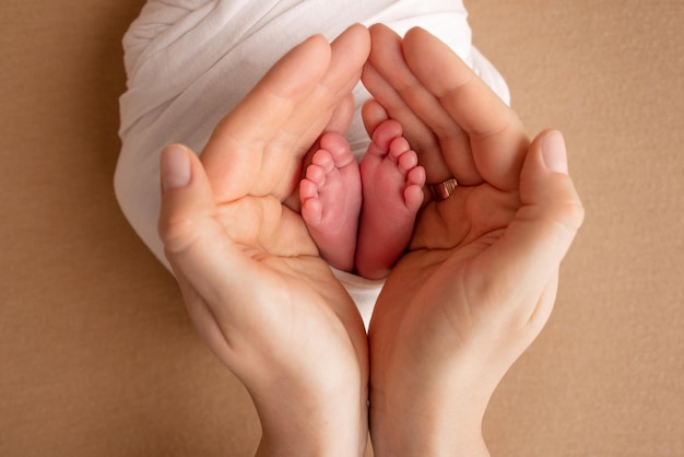 Die Handflächen der Eltern Eine Mutter hält die Füße eines Neugeborenen in einer weißen Decke auf einem beige-orangefarbenen Hintergrund Die Füße eines Neugeborenen in den Händen der Eltern Foto von Fußabsätzen und Zehen