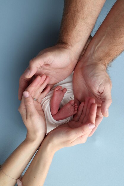 Die Handflächen der Eltern, Vater und Mutter, halten die Beine, die Füße eines Neugeborenen in einer weißen Hülle auf blauem Hintergrund, Füße, Fersen, Fersen und Zehen eines Neuborenen, professionelles Makrofoto