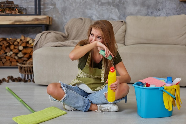 Die Hausfrau fühlt sich müde, wenn sie im Wohnzimmer auf dem Boden sitzt, nachdem sie zu Hause mit einem blauen Eimer gereinigt hat, der mit Chemikalien und Einrichtungen zum Aufräumen gefüllt ist