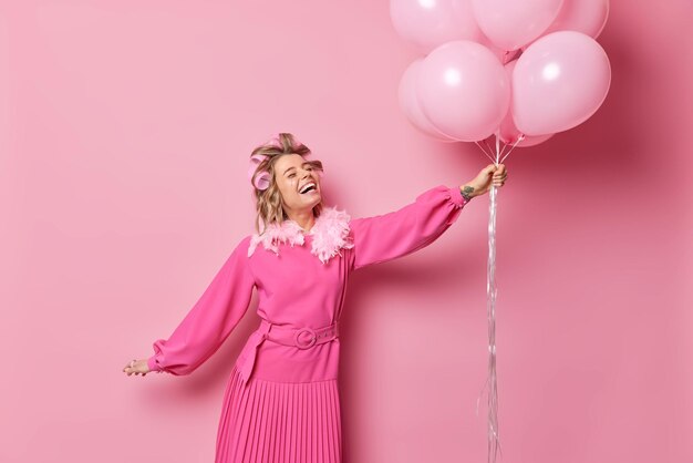 Die horizontale Aufnahme einer fröhlichen Europäerin, die festliche rosafarbene Lockenwickler hat, hat Spaß und genießt Feiertänze mit einem Haufen aufgeblasener Luftballons vor rosigem Hintergrund und hat eine optimistische Stimmung