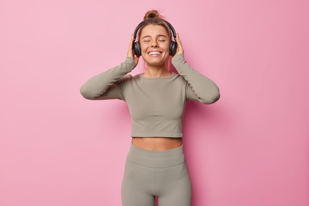 Die horizontale Aufnahme einer glücklichen, schlanken Frau in Activewear hört Musik über Kopfhörer, während das Training im Fitnessstudio die Augen geschlossen hält, isoliert vor rosa Hintergrund. Sportliche Frau genießt Playlist
