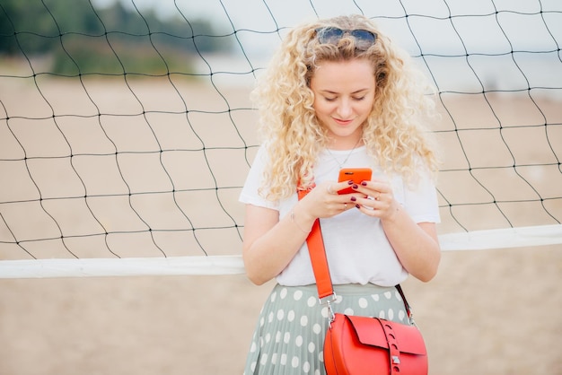 Die horizontale Aufnahme einer gut aussehenden Frau mit lockigem Haar enthält moderne Handy-Textnachrichten an Freunde, die in der Nähe von Tennisnetzspaziergängen am Strand stehen. Schöne Frau surft auf dem Handy im Internet