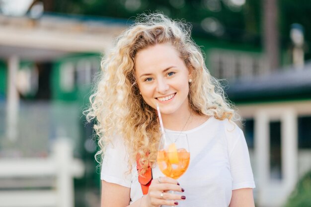 Die horizontale Aufnahme einer hübschen lockigen jungen Frau mit charmantem Lächeln hält ein Glas frischen Orangencocktails und hat einen Spaziergang im Freien vor verschwommenem Hintergrund. Menschen gehen und Freizeitkonzept