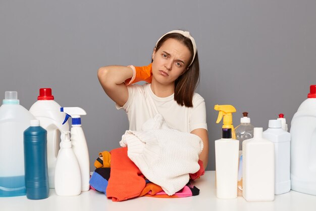 Die horizontale Aufnahme einer müden, erschöpften Frau mit Handschuhen sitzt am Tisch mit vielen Waschmitteln und schmutziger Wäsche, die beim Waschen von Müdigkeit und Schmerzen am Hals isoliert auf grauem Hintergrund posieren muss