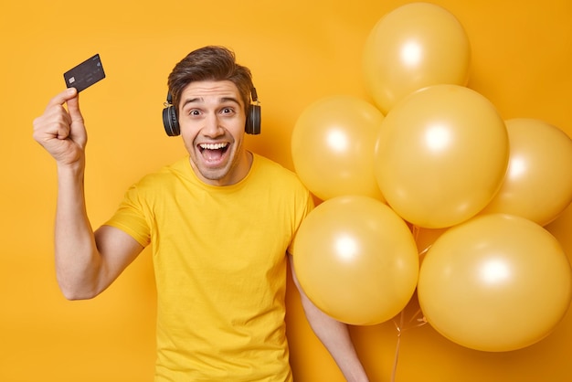 Die horizontale Aufnahme eines fröhlichen jungen Mannes hält eine Plastikkarte und einen Haufen aufgeblasener Luftballons, die fröhlich feiern, den Geburtstag feiern, Geld für die Vorbereitung auf die Party ausgeben, ein lässiges T-Shirt tragen und Musik hören