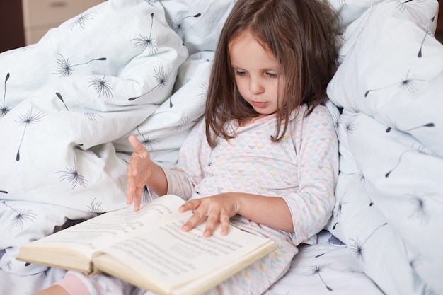 Die horizontale Aufnahme eines hübschen kleinen Mädchens, das ein interessantes Buch in ihrem Zimmer liest, während es im Bett unter einer Decke mit Löwenzahn liegt, sieht konzentriert und ernst aus, niedliches dunkelhaariges weibliches Kind ruht zu Hause.