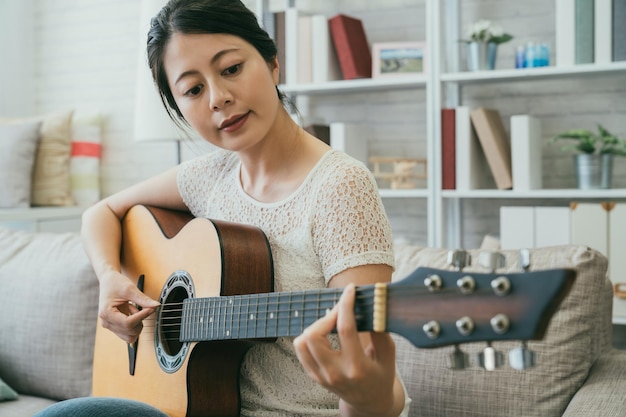 Die hübsche asiatische Japanerin in Freizeitkleidung zupft die Saite, während sie lernt, auf der Couch in einem gemütlichen, hellen Wohnzimmer zu Hause eine Akustikgitarre zu spielen.