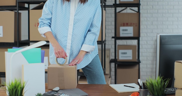 Die Inhaberin eines Online-Shops arbeitet an einem Computer an ihrem Schreibtisch in einer Lagerhalle. Eine Mitarbeiterin packt einen gelben Pullover in einem Raum mit Regalen voller Pakete