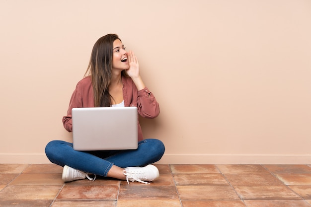 Die Jugendlichstudentenfrau, die auf dem Boden mit einem Laptop schreit mit dem breiten Mund sitzt, öffnen sich zur Seite