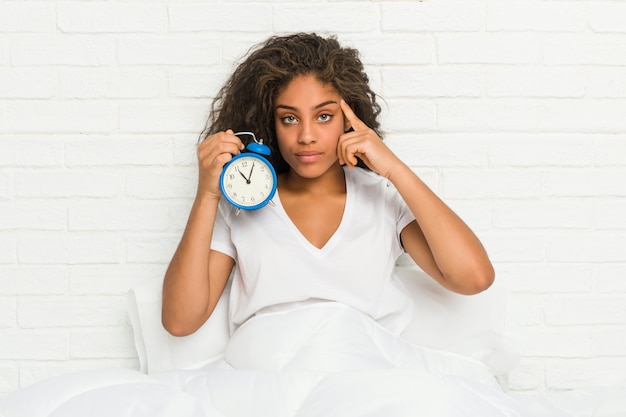 Die junge Afroamerikanerfrau, die auf dem Bett hält einen Wecker zeigt seinen Tempel mit dem Finger sitzt und denkt, konzentrierte sich auf eine Aufgabe.