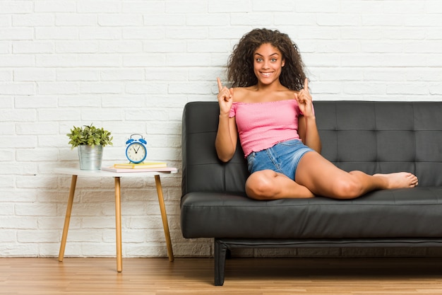 Die junge Afroamerikanerfrau, die auf dem Sofa sitzt, zeigt mit beiden vorderen Fingern an, die sich eine Leerstelle zeigen.