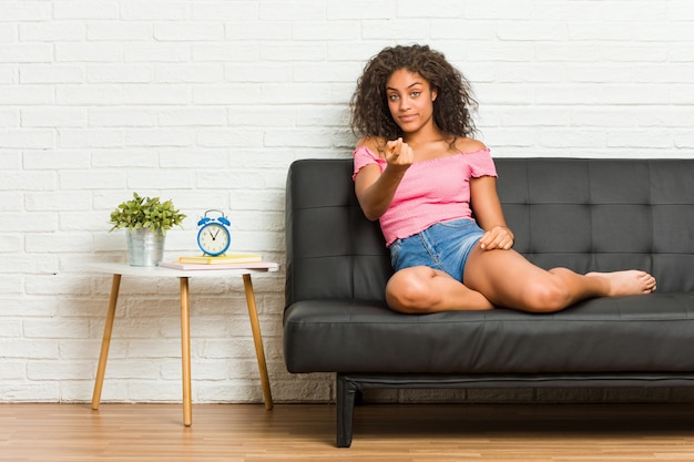 Die junge Afroamerikanerfrau, die auf dem Sofa zeigt mit dem Finger auf Sie sitzt, als ob Einladung näher kommen.