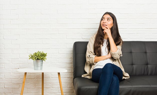 Die junge arabische Frau, die auf dem Sofa sitzt, entspannte sich das Denken an etwas, das einen Kopienraum betrachtet.