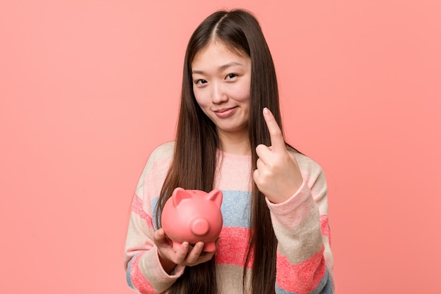 Die junge asiatische Frau, die ein Sparschwein zeigt mit dem Finger auf Sie hält, als ob Einladung näher kommen.