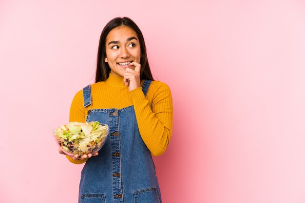 Die junge asiatische Frau, die einen Salat hält, lokalisierte das entspannte Denken an etwas, das einen Kopienraum betrachtet.