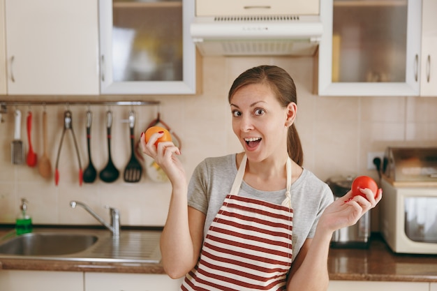 Die junge attraktive Frau mit Schürze entscheidet sich für eine rote oder gelbe Tomate in der Küche. Diätkonzept. Gesunder Lebensstil. Kochen zu Hause. Essen zubereiten.