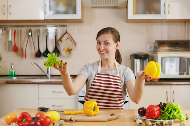 Die junge attraktive lächelnde Frau in einer Schürze mit Kopfsalatblatt und gelbem Pfeffer in der Küche. Diätkonzept. Gesunder Lebensstil. Kochen zu Hause. Essen zubereiten.