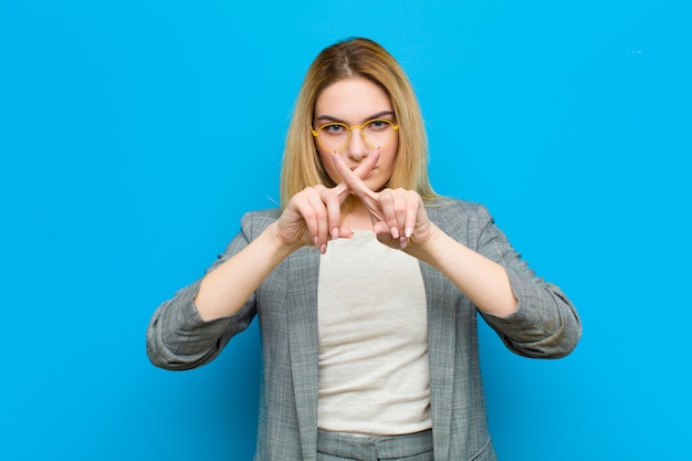 Die junge blonde Frau, die ernst und mit beiden Fingern missfallen schaut, kreuzte ehrlich in der Ablehnung und bat um Ruhe auf blauer Wand