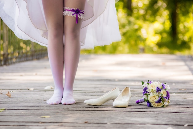 Die junge Braut in einem weißen Kleid, die barfuß auf einer Holzbrücke steht. Schuhe Mädchen und ein Blumenstrauß liegen dort. Braut steht barfuß auf der Brücke.