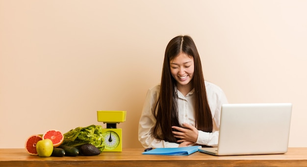 Die junge chinesische Ernährungswissenschaftlerin, die mit ihrem Laptop arbeitet, lacht glücklich und hat Spaß, Hände auf Bauch zu halten.