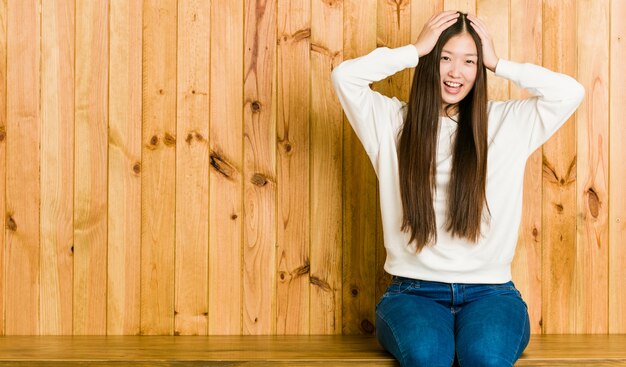 Die junge chinesische Frau, die auf einem hölzernen Platz sitzt, lacht, Hände auf Kopf froh halten. Glück-Konzept.