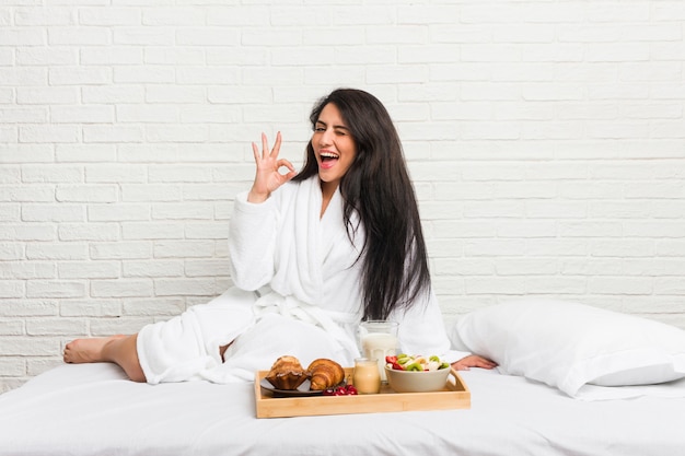 Die junge curvy Frau, die ein Frühstück auf dem Bett nimmt, blinzelt ein Auge und hält eine okaygeste mit der Hand.