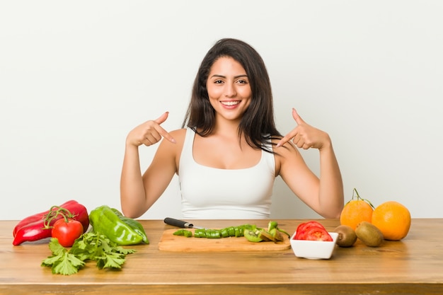 Die junge curvy Frau, die eine gesunde Mahlzeit vorbereitet, zeigt unten mit den Fingern, positives Gefühl.