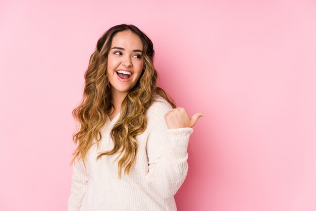 Die junge curvy Frau, die in einer rosa Wand aufwirft, lokalisierte Punkte mit dem Daumenfinger weg, lachte und sorglos.